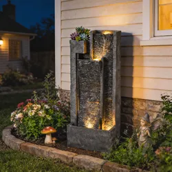 A lighted tiered garden fountain with cascading water, surrounded by flowers, a mushroom ornament, and a fairy statue, creating a decorative outdoor scene.