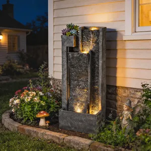A lighted tiered garden fountain with cascading water, surrounded by flowers, a mushroom ornament, and a fairy statue, creating a decorative outdoor scene.