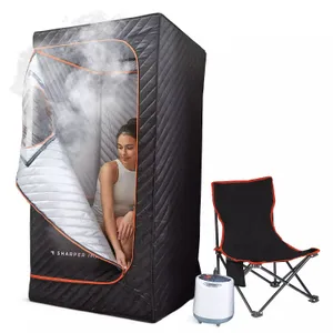 A portable sauna tent with a quilted black exterior and a zippered opening, containing a steam generator and accompanied by a black folding chair with orange trim.