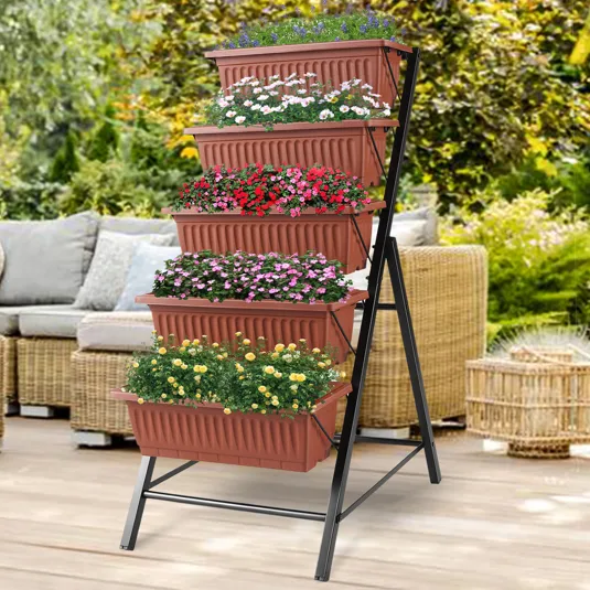 A tiered plant stand features five terracotta-colored rectangular planters stacked vertically, each filled with different colorful flowers, supported by a black metal frame. It's set on a patio with wicker furniture in the background.