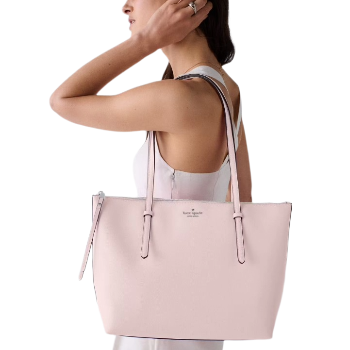 A person holds a light pink Kate Spade New York tote bag with long straps and a minimalist design, suitable for carrying daily essentials.