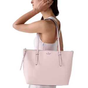 A person holds a light pink Kate Spade New York tote bag with long straps and a minimalist design, suitable for carrying daily essentials.
