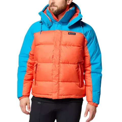 A man wears a two-tone puffer jacket with an orange body and blue shoulders, featuring a hood and black pants.