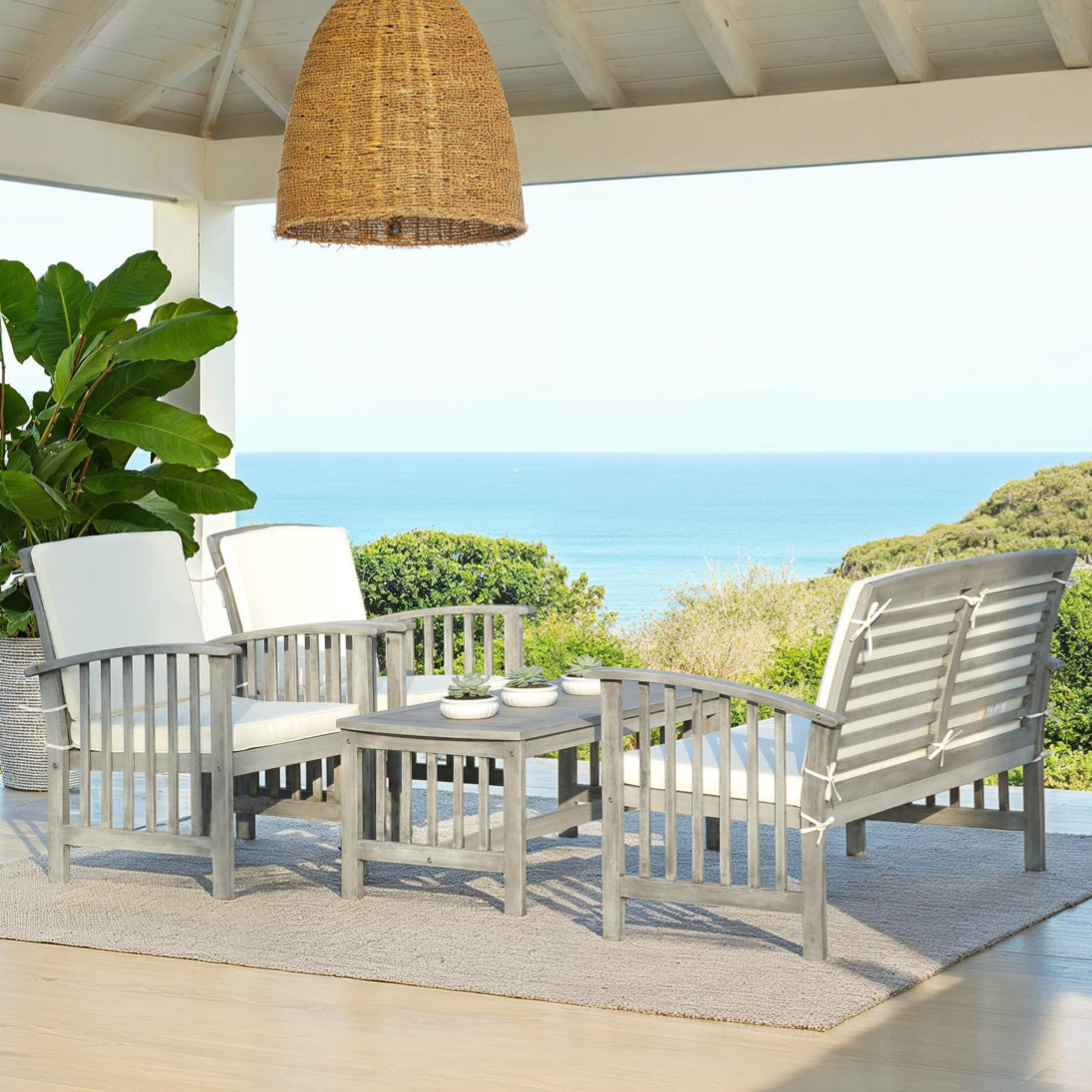Outdoor patio set featuring two armchairs, a loveseat, and a coffee table, all in light wood with white cushions. Positioned on a woven rug under a wicker pendant light with a coastal backdrop.