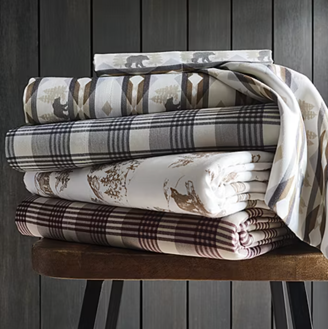 Stack of patterned bedsheets featuring bear motifs, plaid designs, and nature-themed prints in earth tones, neatly folded on a wooden bench.