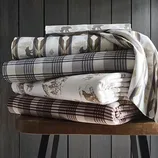 Stack of patterned bedsheets featuring bear motifs, plaid designs, and nature-themed prints in earth tones, neatly folded on a wooden bench.