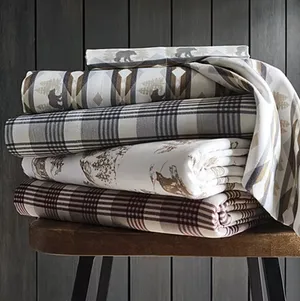 Stack of patterned bedsheets featuring bear motifs, plaid designs, and nature-themed prints in earth tones, neatly folded on a wooden bench.