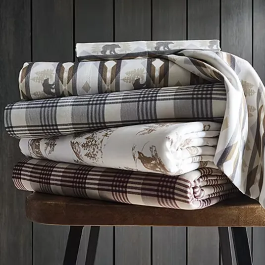 Stack of patterned bedsheets featuring bear motifs, plaid designs, and nature-themed prints in earth tones, neatly folded on a wooden bench.