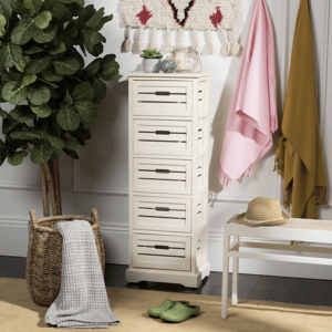 A white five-drawer storage cabinet is shown beside a large leafy plant. Nearby are a woven basket with a gray blanket, a white bench with a straw hat, green shoes, and pink and mustard throws on hooks above.