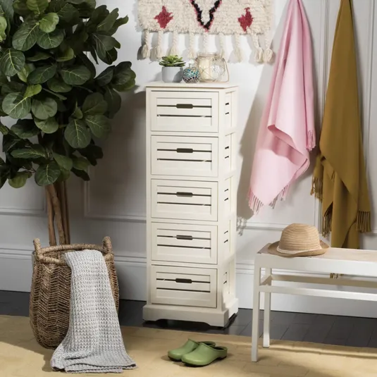 A white five-drawer storage cabinet is shown beside a large leafy plant. Nearby are a woven basket with a gray blanket, a white bench with a straw hat, green shoes, and pink and mustard throws on hooks above.