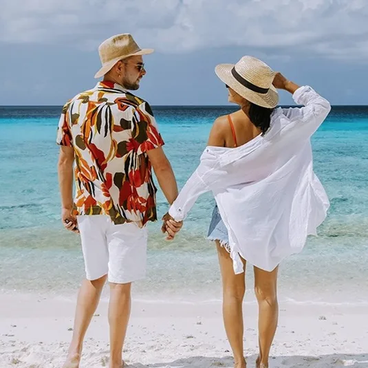 A couple at the beach wearing straw hats, a floral print shirt, white shorts, a white cover-up, and denim shorts, standing on white sand near clear blue water.