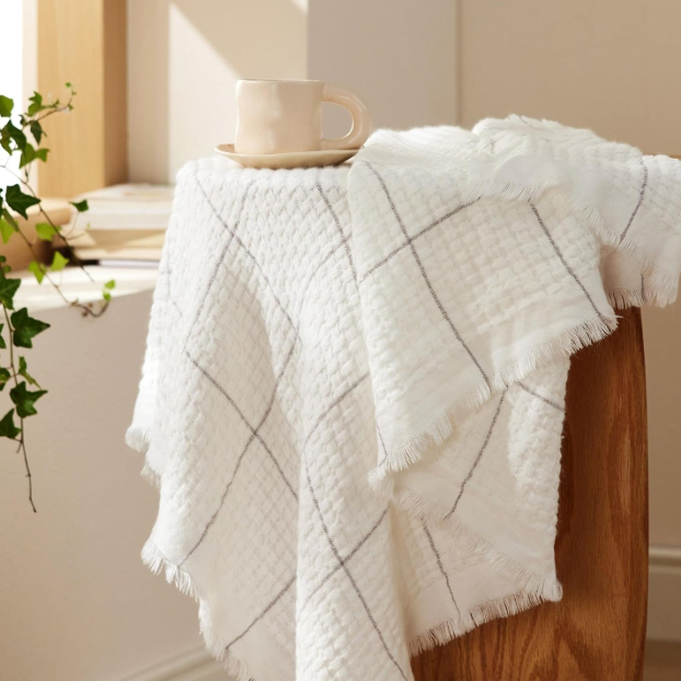 A white throw blanket with a subtle textured pattern and light plaid lines, draped over a wooden surface, accompanied by a light-colored mug.