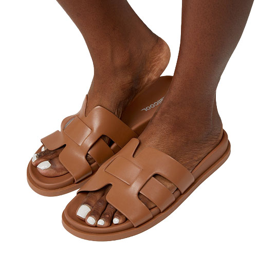 Brown slip-on sandals with interlocking strap design are worn by a person with white toenails.