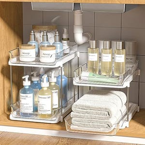 Two tiered, sliding clear plastic cabinet organizers are shown storing various bottles and towels under a sink.