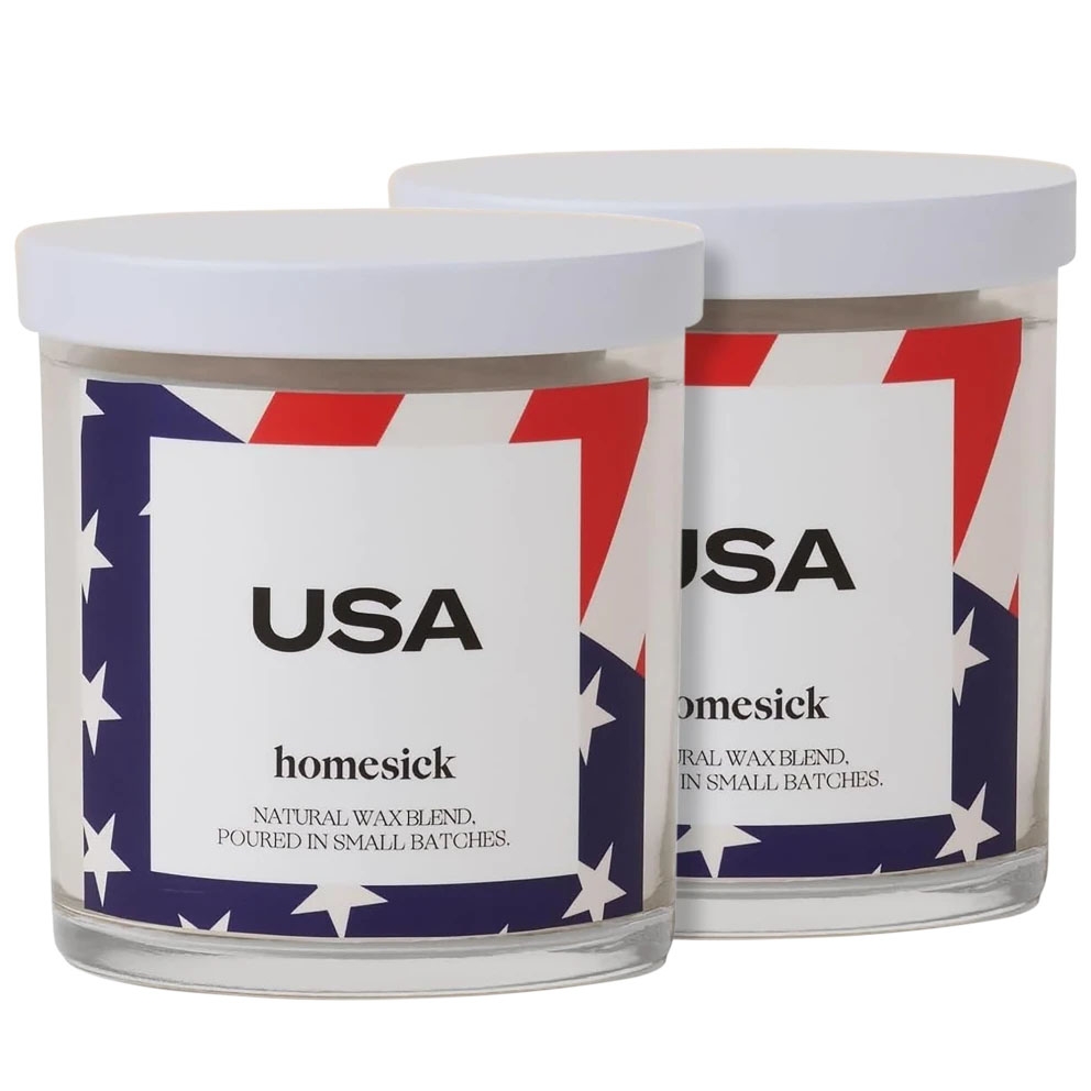 Two candles in glass jars with white lids featuring \“USA\“ and \“homesick\“ on the labels. The design includes a red, white, and blue star pattern.