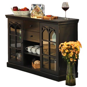 Black buffet sideboard with glass doors, three drawers and a vase of flowers, accompanied by a fruit basket, a decorative plate, and a glass of wine on top.