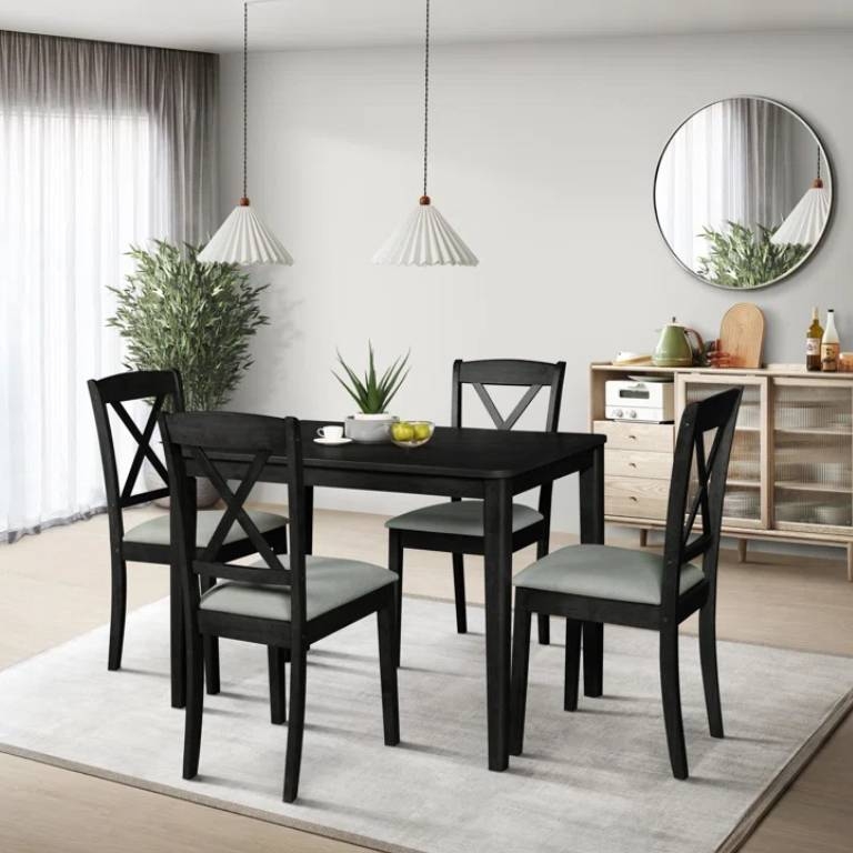 Dining set with a black table and four chairs on a white rug, featuring cross-back designs. Two pendant lights hang above, and there's a sideboard with decorative items against the wall. A round mirror and potted plant complete the room decor.