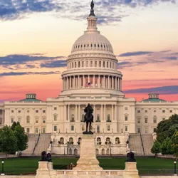The U.S. Capitol building at sunset, featuring its iconic dome and an equestrian statue in the foreground, with a U.S. flag displayed prominently.
