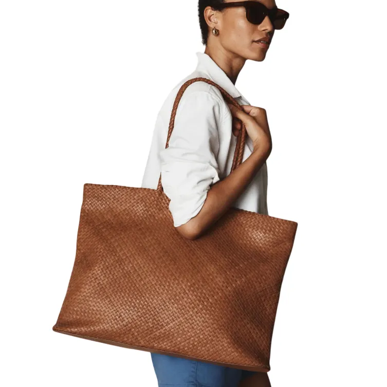 A person wearing a white shirt and sunglasses carries a large, woven brown tote bag on their shoulder.