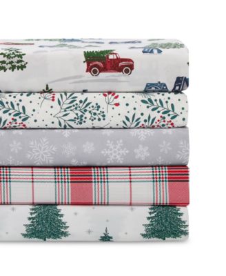 A stack of four queen-sized holiday-themed sheet sets featuring patterns with a red truck, snowflakes, Christmas trees, and plaid design in red, green, white, and grey colors.