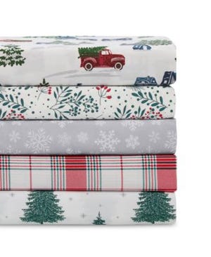 A stack of four queen-sized holiday-themed sheet sets featuring patterns with a red truck, snowflakes, Christmas trees, and plaid design in red, green, white, and grey colors.