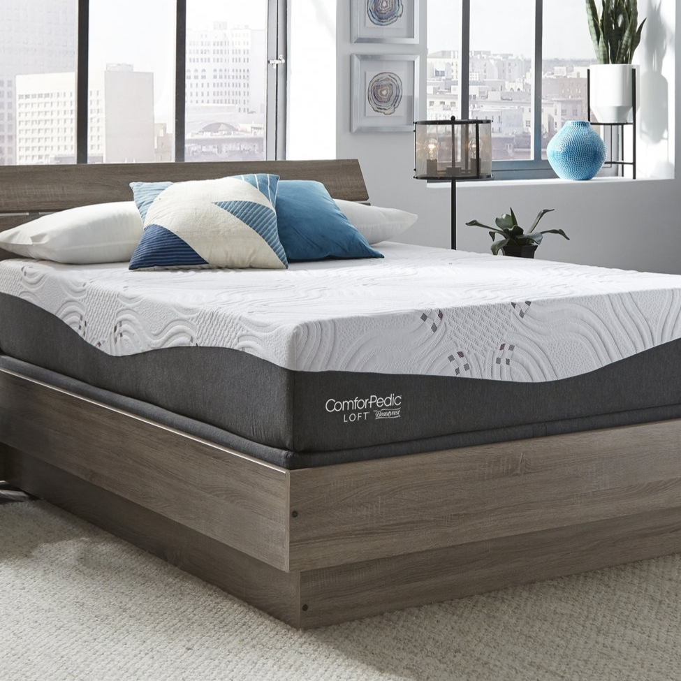 A modern bedroom with a ComforPedic Loft mattress on a wooden platform bed, accented with blue and geometric pillows. Decor includes a lantern-style lamp, potted plant, and city view through large windows.