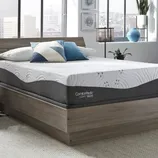 A modern bedroom with a ComforPedic Loft mattress on a wooden platform bed, accented with blue and geometric pillows. Decor includes a lantern-style lamp, potted plant, and city view through large windows.