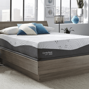 A modern bedroom with a ComforPedic Loft mattress on a wooden platform bed, accented with blue and geometric pillows. Decor includes a lantern-style lamp, potted plant, and city view through large windows.