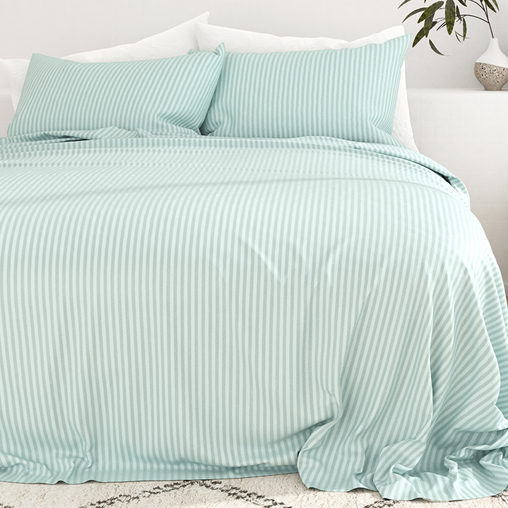 A bed is covered with light blue striped bedding, including matching pillows and a duvet. A plant is placed on a nearby nightstand.