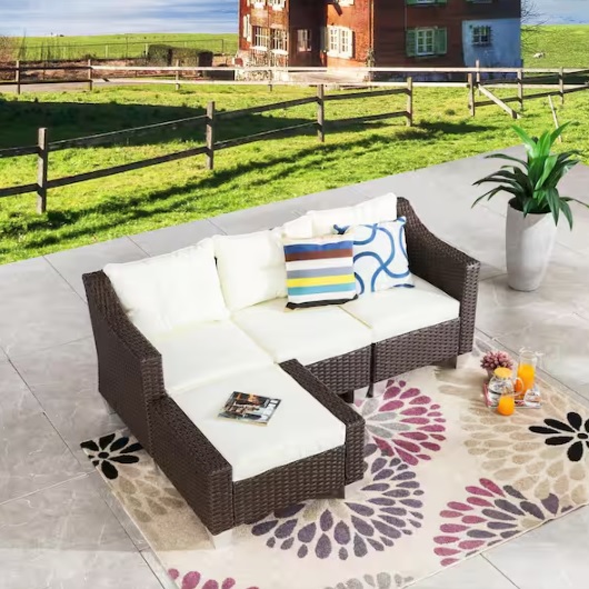 Outdoor rattan sectional sofa with white cushions and a colorful pillow on a floral patterned rug, accompanied by potted plant and refreshments on the floor.