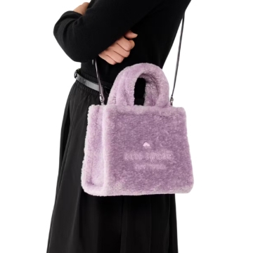 A person holds a fluffy, light purple handbag with a shoulder strap, featuring the words \“the tote bag\“ embroidered on its side.