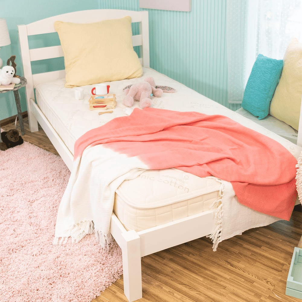 A child's bedroom features a white bed with a plush mattress, soft pillows, a pink and white throw, and a plush toy. A small side table holds a stuffed animal, with a pink rug on the wooden floor.