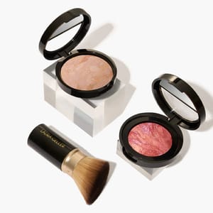 Two compact cases of baked blushes in light and pink shades, accompanied by a makeup brush with a gold ferrule and \“Laura Geller\“ branding on the handle.