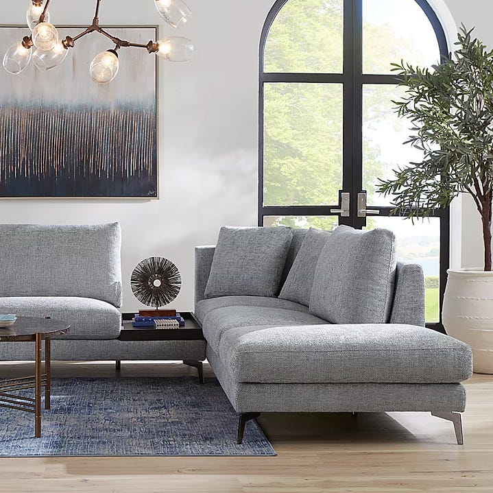 Living room setup with a light gray sectional sofa, a rectangular coffee table, a blue patterned rug, and a wall painting. A decorative piece sits on the coffee table, with a modern chandelier overhead and a potted plant near the arched window.