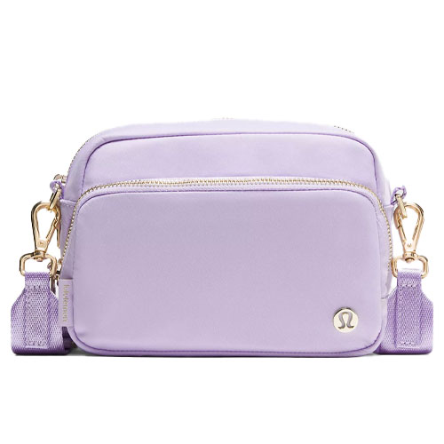 A light purple crossbody bag with gold hardware, featuring two zippered compartments and an adjustable strap with clips. It has a small circular emblem on the front pocket.