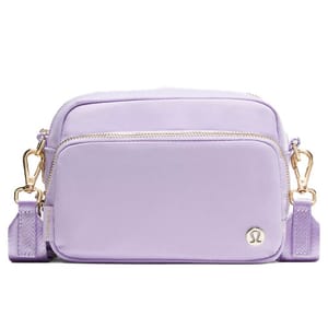 A light purple crossbody bag with gold hardware, featuring two zippered compartments and an adjustable strap with clips. It has a small circular emblem on the front pocket.