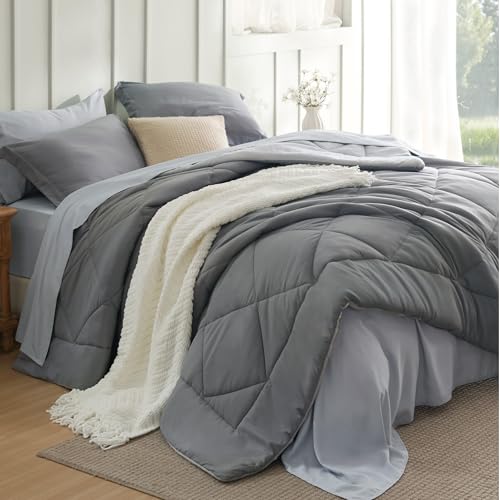 A neatly made bed with a grey comforter, matching grey pillows, and a white textured blanket draped over the edge. A beige pillow contrasts against the grey bedding, set in a bright, well-lit room with a nightstand.