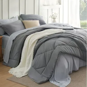 A neatly made bed with a grey comforter, matching grey pillows, and a white textured blanket draped over the edge. A beige pillow contrasts against the grey bedding, set in a bright, well-lit room with a nightstand.
