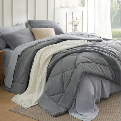 A neatly made bed with a grey comforter, matching grey pillows, and a white textured blanket draped over the edge. A beige pillow contrasts against the grey bedding, set in a bright, well-lit room with a nightstand.