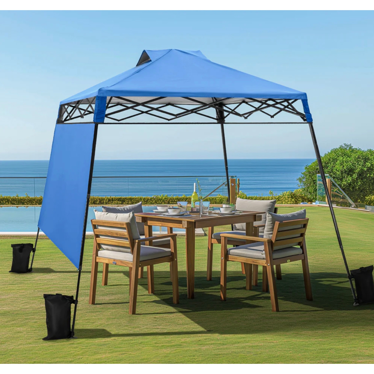 An 8' x 8' blue pop-up canopy provides shade over a wooden outdoor dining set with four cushioned chairs on a grassy lawn, with a pool and ocean visible in the background.