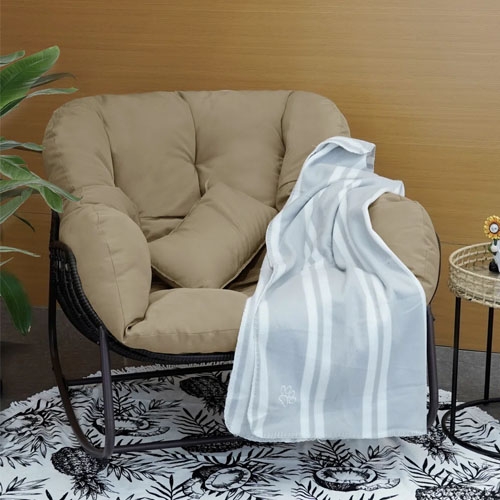 A beige cushioned chair with a light grey and white striped blanket draped over the arm, placed on a patterned rug next to a small round woven table.