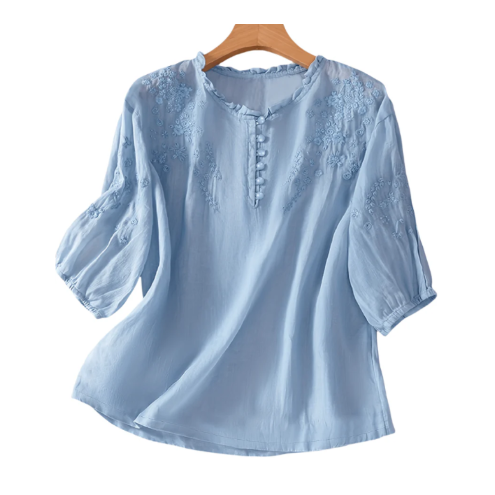 Light blue blouse with floral embroidery on the chest and sleeves, featuring a round neckline with button details and three-quarter length sleeves.