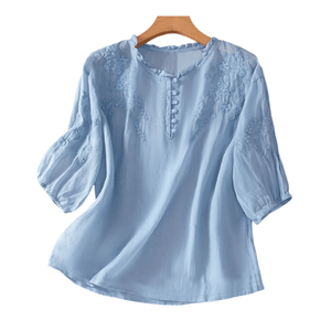 Light blue blouse with floral embroidery on the chest and sleeves, featuring a round neckline with button details and three-quarter length sleeves.