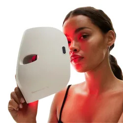 A woman holds a white TherFace Mask Glo, a skincare device emitting red light, designed for facial treatment.