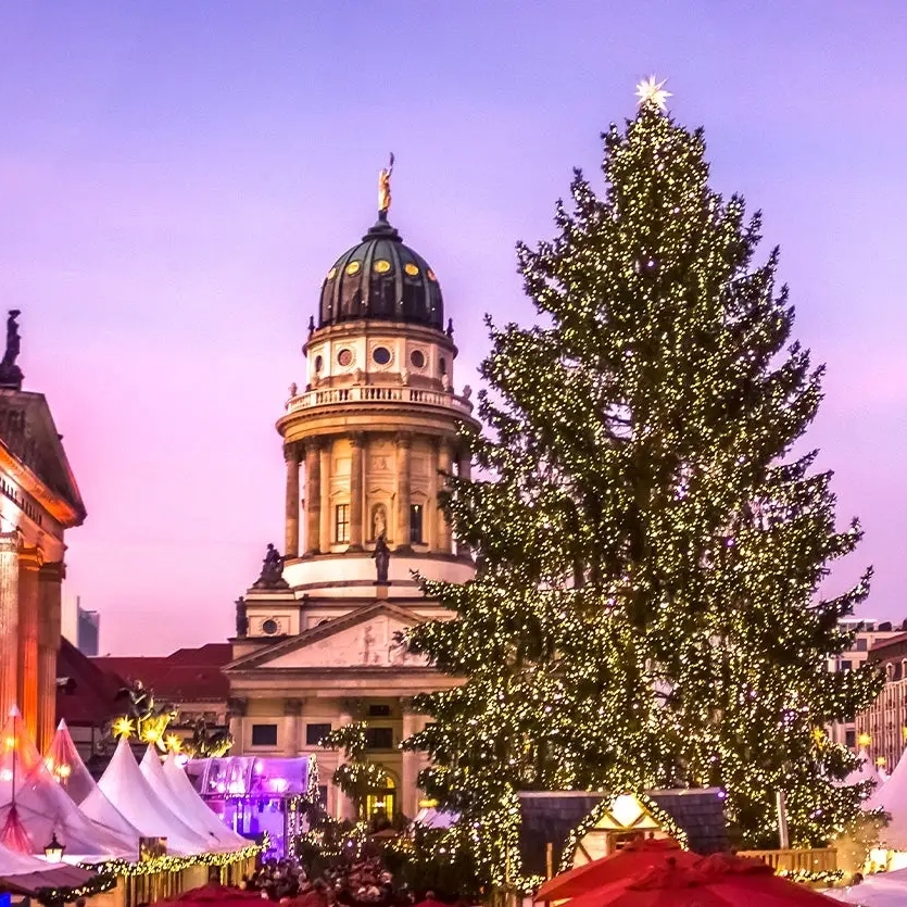 A decorated Christmas tree and festive market stalls set against the backdrop of a neoclassical building at dusk.