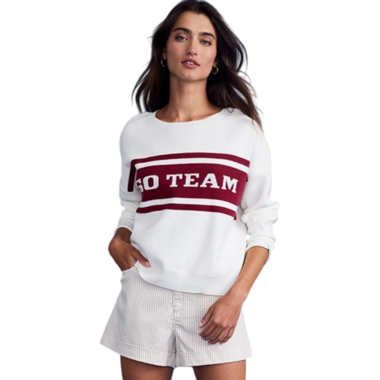 A woman wears a white sweater with \“GO TEAM\“ in red, paired with white striped shorts and white high-top sneakers. She poses against a blue backdrop.