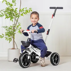 A child rides a white tricycle with a push handle, next to a potted plant. The tricycle features black wheels and a black basket.