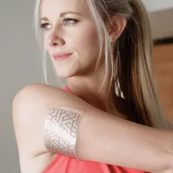 A woman stretches her arm while wearing a patterned adhesive patch labeled \“Kino\“ on her upper arm.