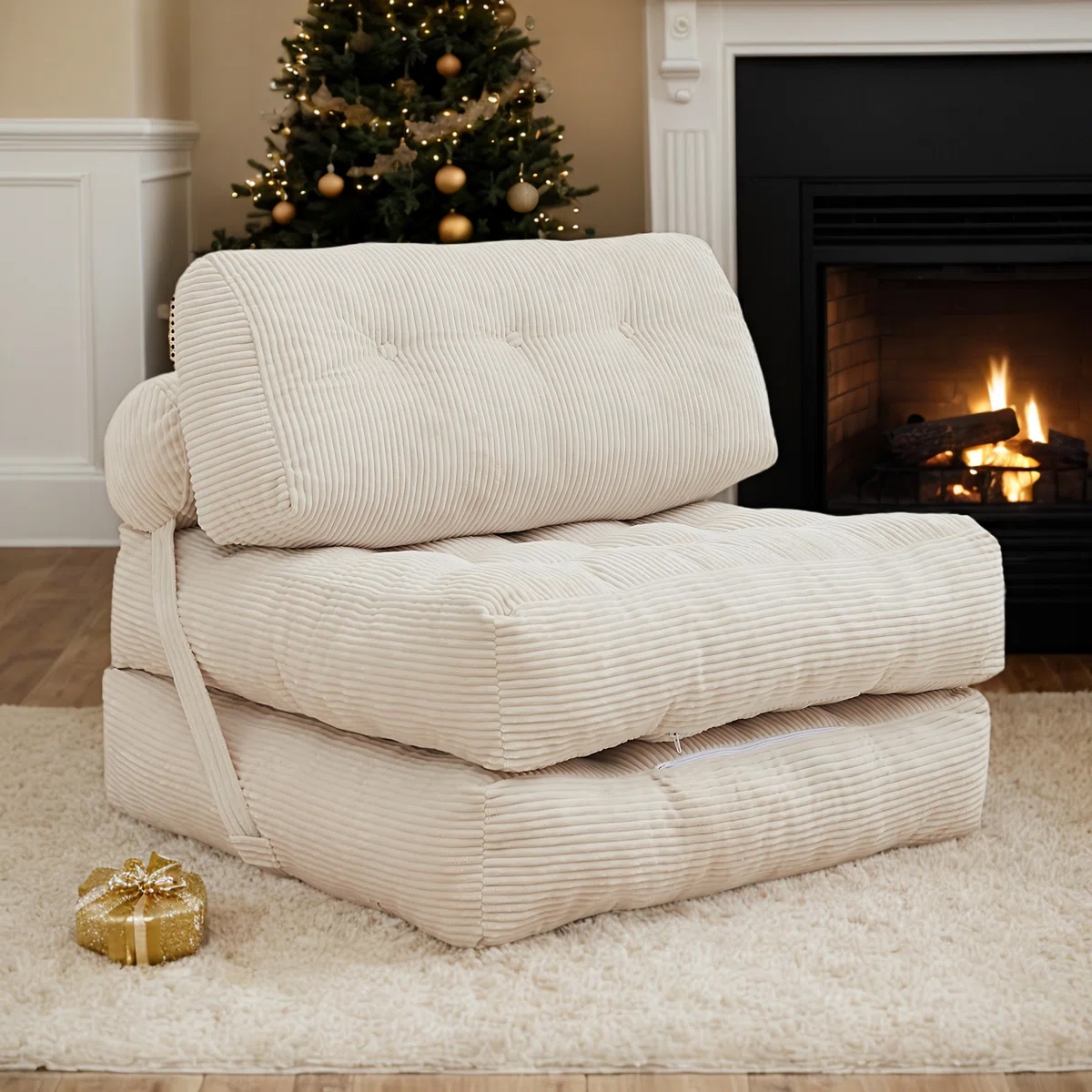 A cream-colored, tufted convertible sleeper chair is shown in front of a fireplace, with a Christmas tree and gift in the background.