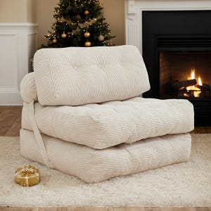 A cream-colored, tufted convertible sleeper chair is shown in front of a fireplace, with a Christmas tree and gift in the background.